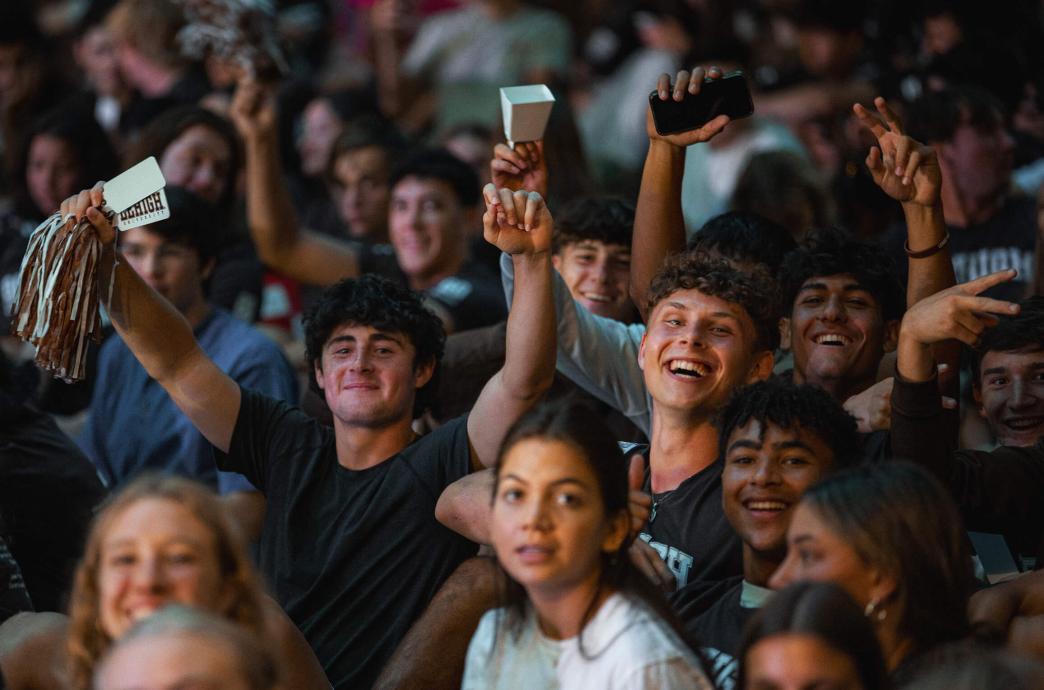 A crowd of students sitting in the grass, smiling, a few with their arms raised holding pompoms and cowbells.