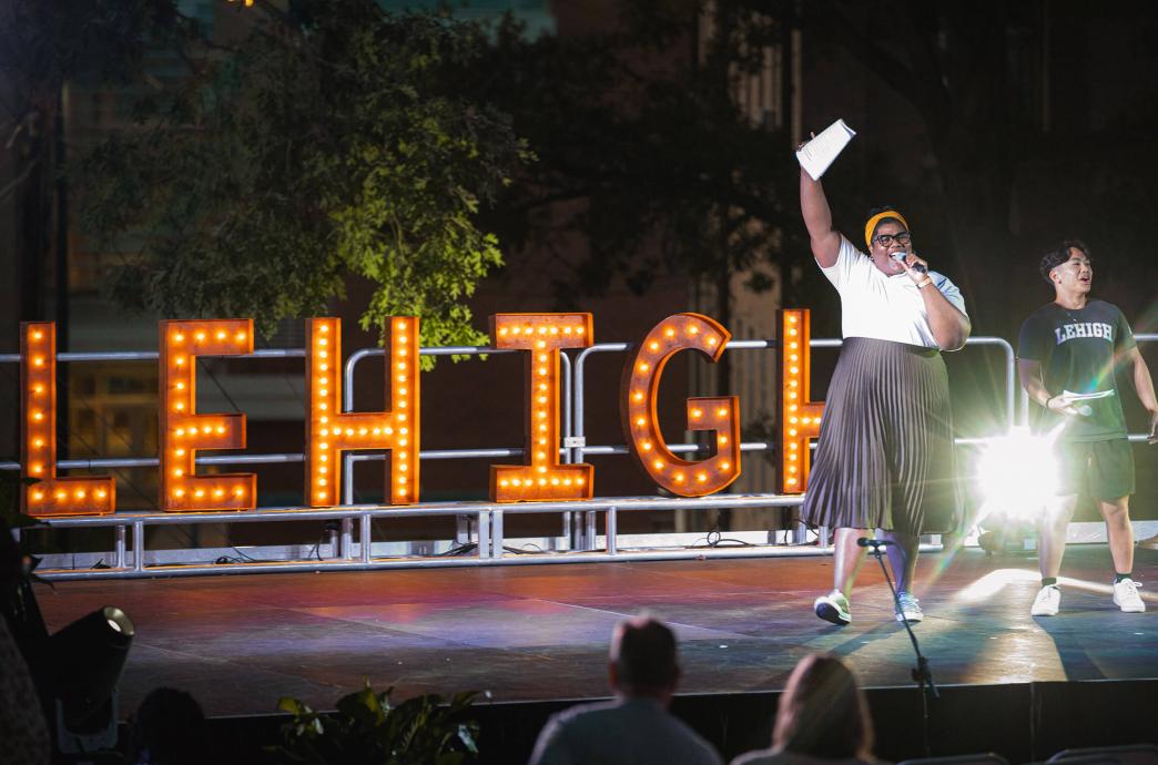 A woman in a long skirt stands on a stage beside a student in a brown Lehigh shirt before illuminated "Lehigh" marquee letters.