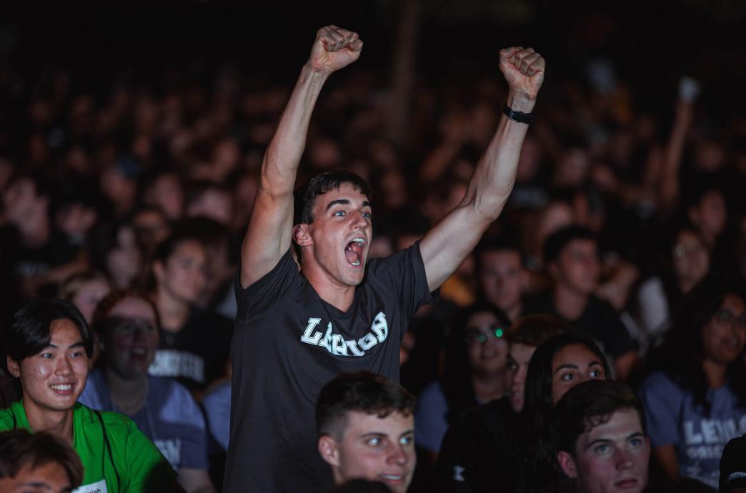 A student wearing a Lehigh shirt stands with his arms raised above the crowd, cheering.