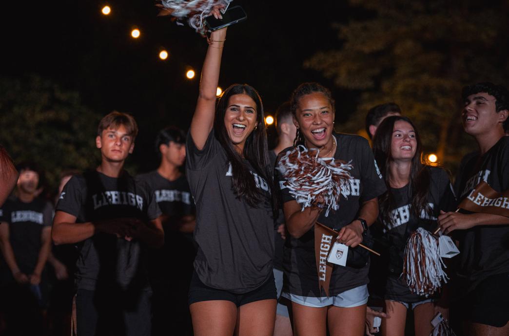 Two students stand in the center of a crowd yelling in celebration, waving pompoms and pennants.