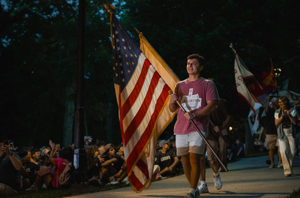 A student wearing a Student Alumni shirt walks past seated students, carrying a large American flag and a smile.