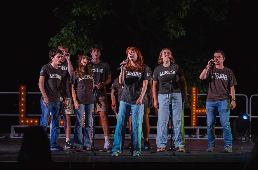 An acapella group wears brown Lehigh shirts on a stage before a Lehigh marquee, singing into microphones.