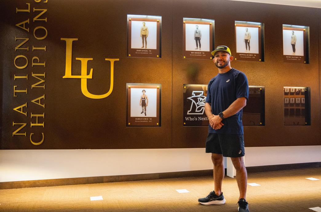 Darian Cruz stands against a wall of photos showing his picture alongside other Lehigh wrestling champions.