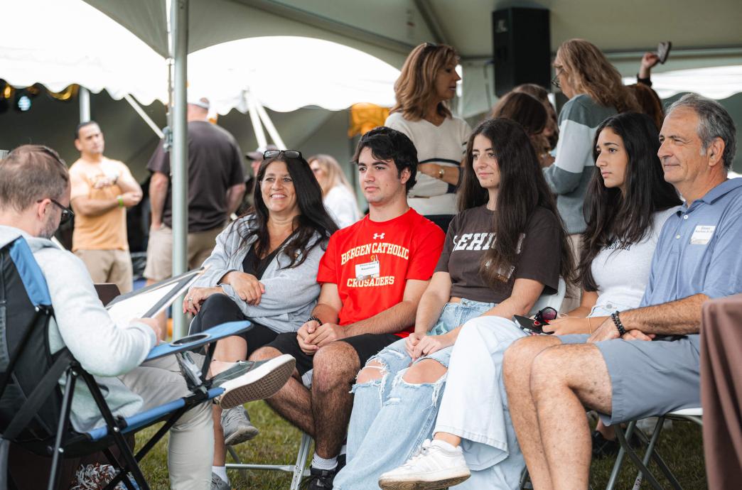 Five family members sit in folding chairs for a caricature artist under a large white tent.