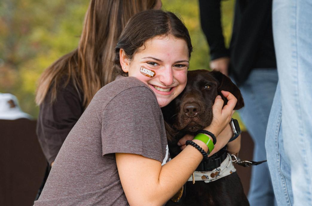 A girl wearing a Lehigh sticker on her face snuggles close to a chocolate labrador retriever.