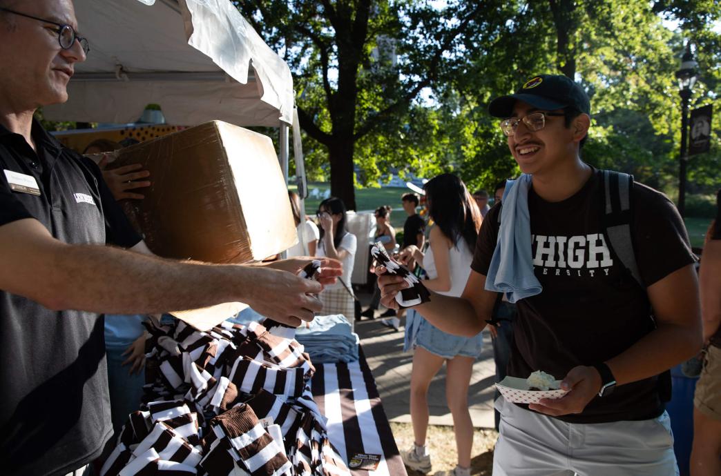 A staff member in a Lehigh shirt stands behind a pile of brown and white striped socks, handing a pair to a man wearing a brown Lehigh shirt.