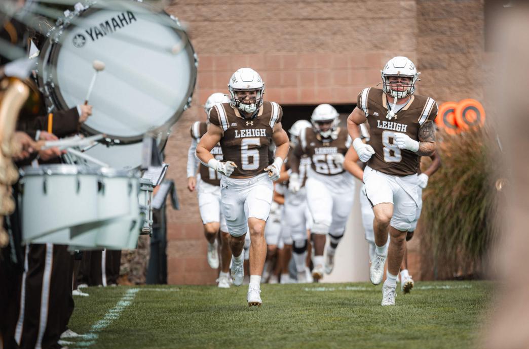 Football players in full game gear enter the field, running down a row flanked by the marching band.