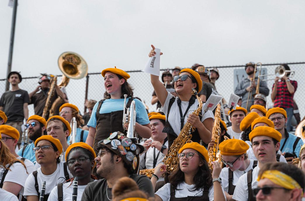 Students holding instruments and wearing yellow berets cheer in the stands of a football game.