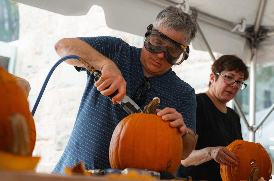 A man wearing safety goggles uses a power tool to carve the top off of a pumpkin.