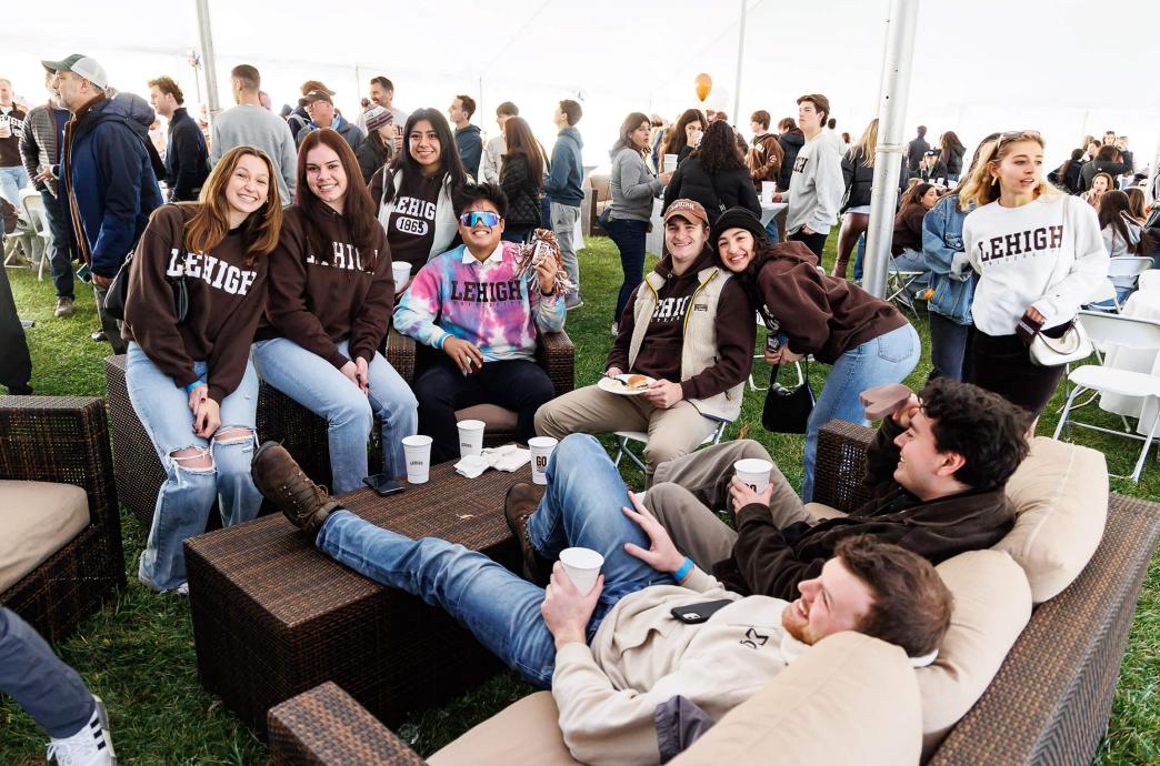 A group of alumni sit on comfortable, plush outdoor sofas ad smile for the camera.
