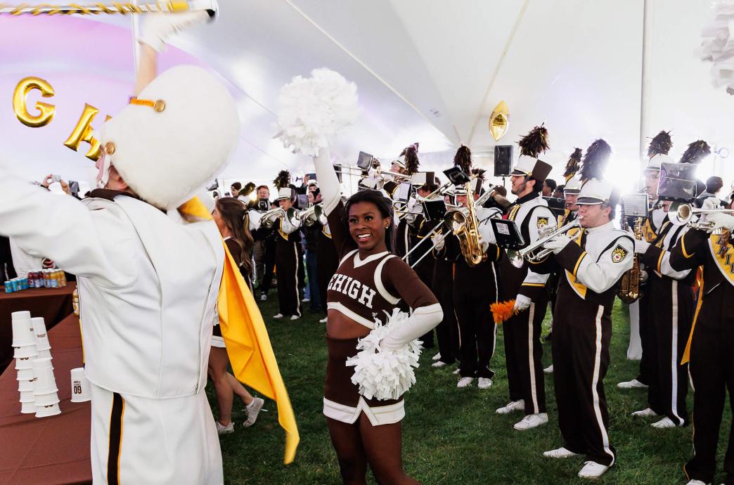 A cheerleader and marching band perform under a white party tent.