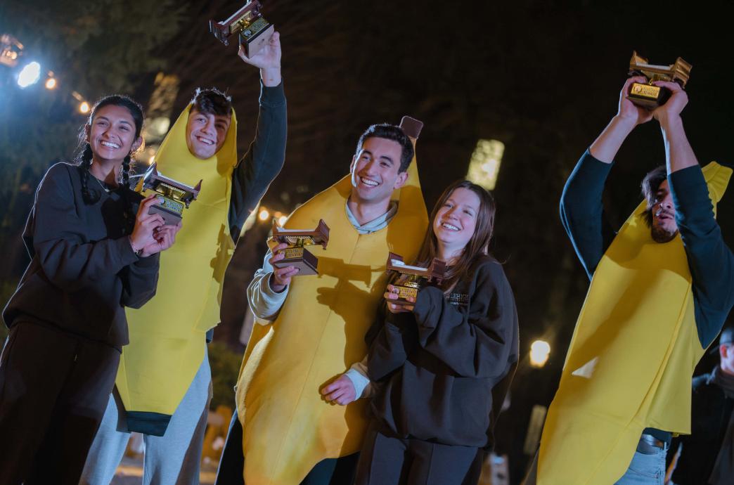 A team of five people, two people wearing all black, and three people wearing banana costumes, hold their bed race trophies.