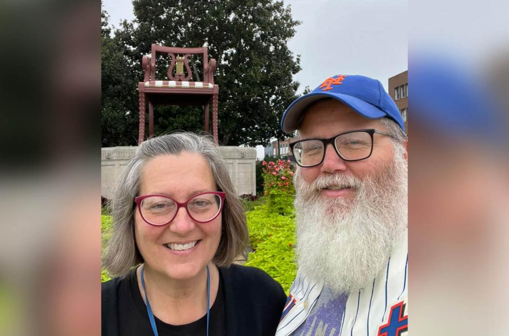 Woman wearing red glasses stands outdoors beside her husband who has glasses, a beard, and blue baseball hat.