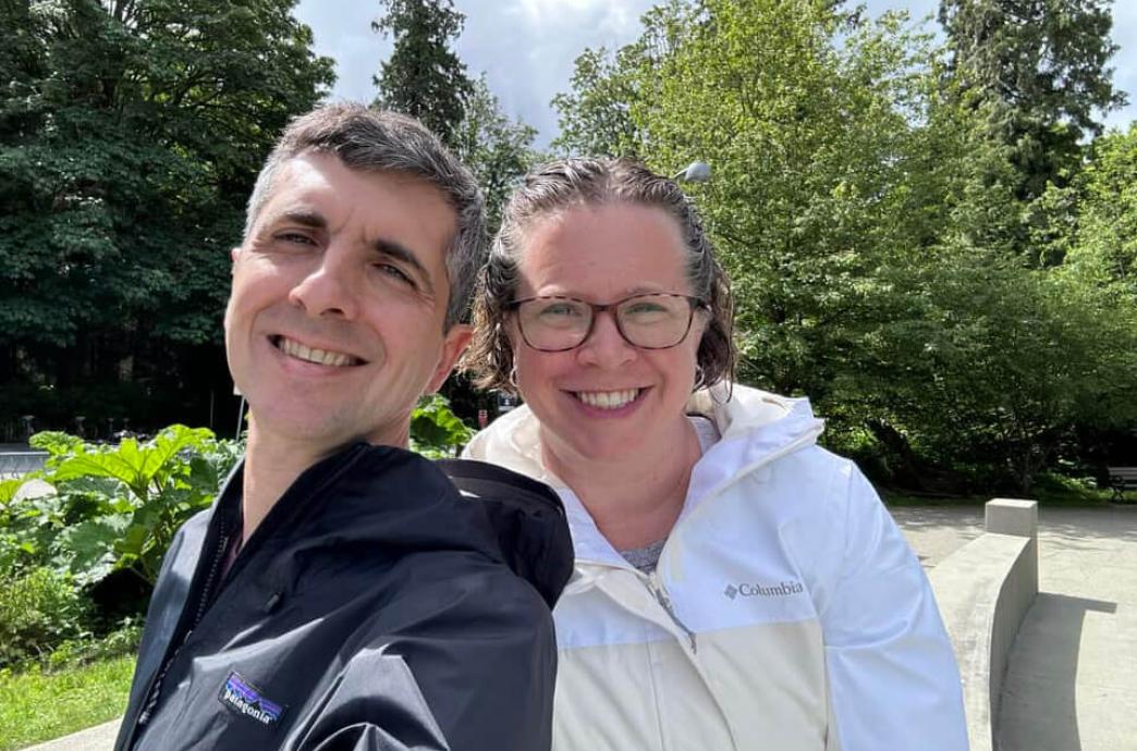 A man and woman smile for the camera outdoors, with the woman positioned slightly behind the man with their heads close together.