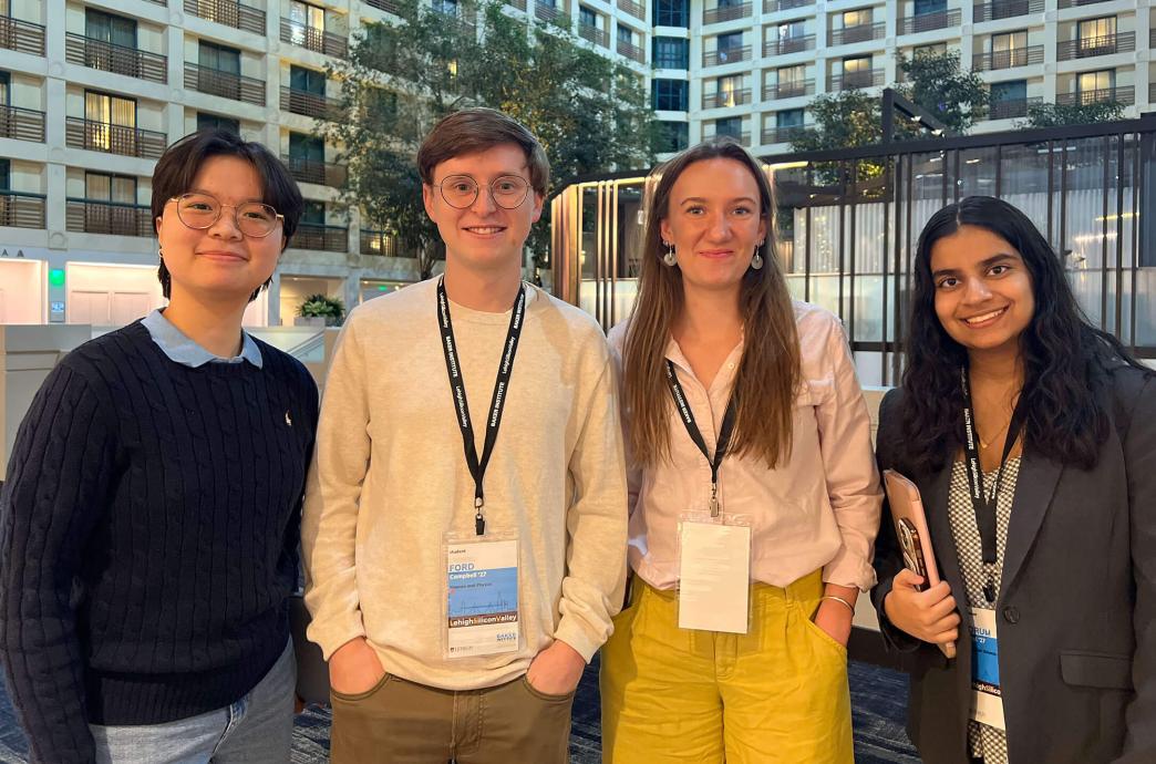 Four students wearing lanyards pose with smiles in front of a tall hotel with balcony suites.