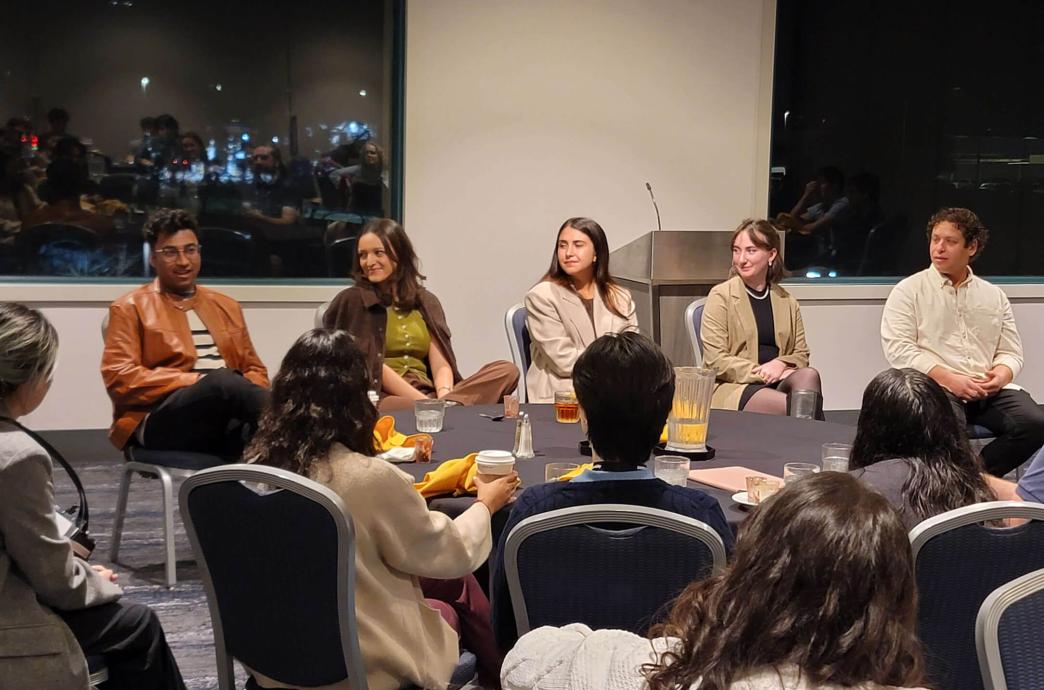 A panel of 5 young alumni sit in chairs in front of a room of students.