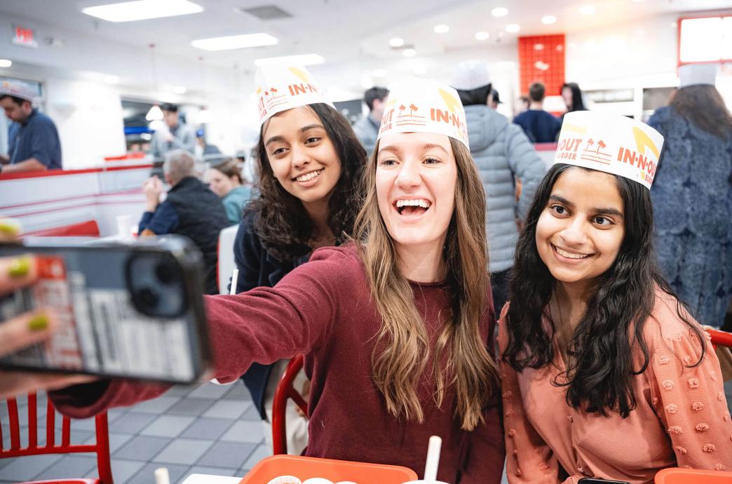 Three female students pose for a selfie in an In-N-Out restaurant with paper hats on their heads.