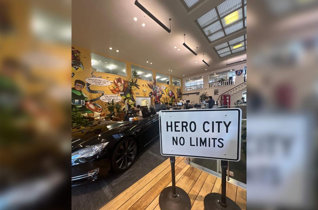 A large white street sign reading “Hero City No Limits” stands in front of a super hero themed room with murals on the walls and a desk made from a Tesla.
