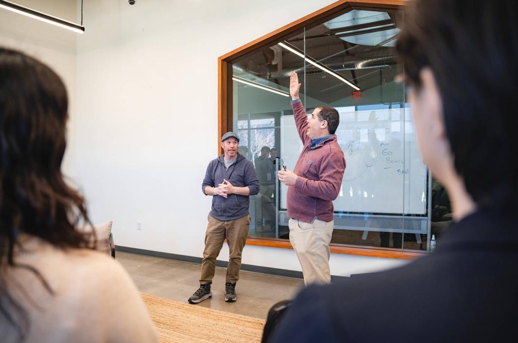A man stands beside a colleague while demonstrating to a group of students in front of a white board while reaching one hand far into the air.