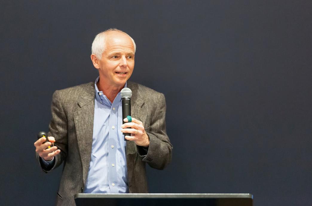 Marc Tarpenning holds a presentation clicker in one hand and a microphone in the other, speaking in front of a black wall to an audience that is not pictured.