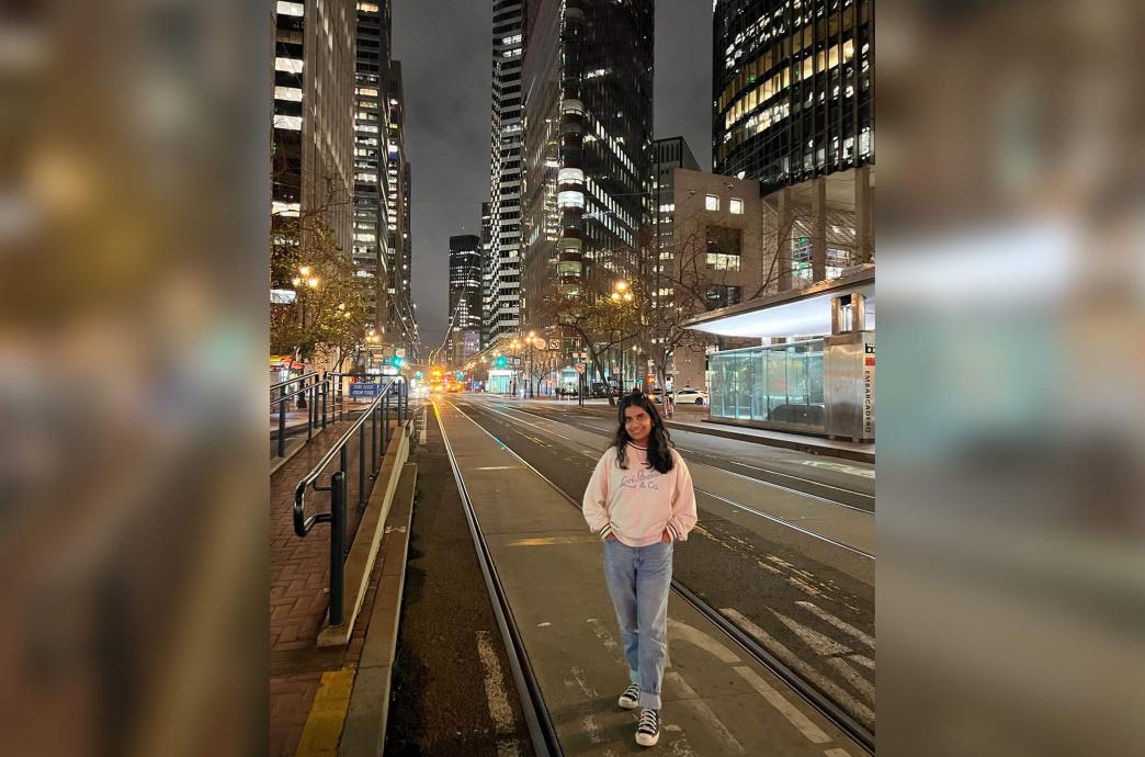 Forum Patel stands on the street car tracks in San Francisco during the night wearing a white sweatshirt and jeans in front of the lit skyline.