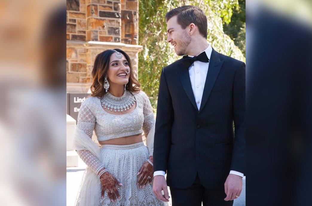 A man in a black tuxedo and a woman in her southeast Asian bridal gown and jewelry smile at each other on their wedding day.