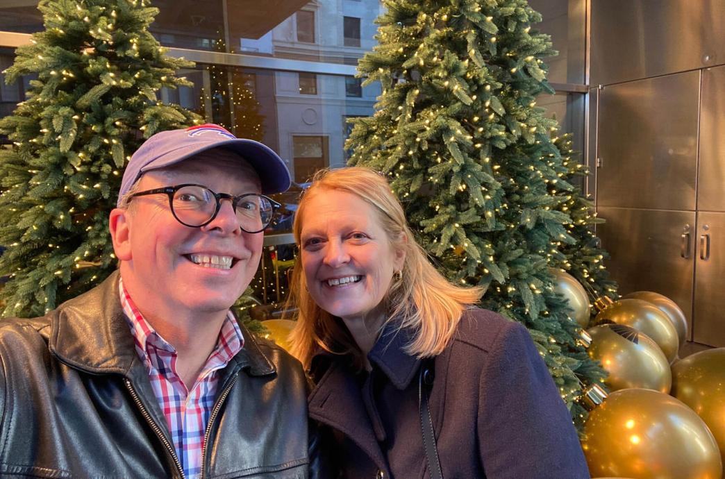 A man and woman pose as the husband takes a selfie in front of evergreen trees and large gold ornaments on the ground.