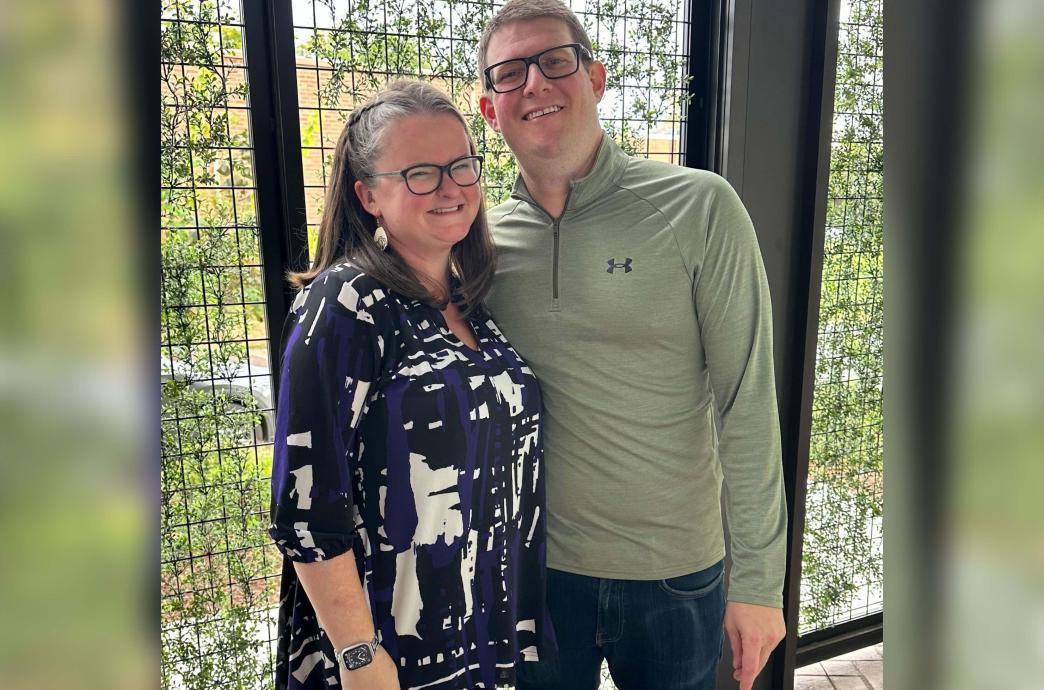 A woman stands for a photo with her husband at her side in front of a tall gated wall.