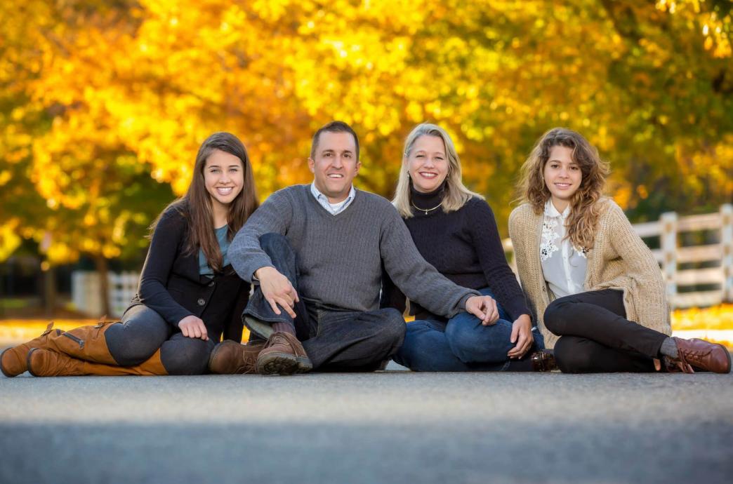 A family of four sits on the asphalt for a portrait with a gold foliage scene behind them.