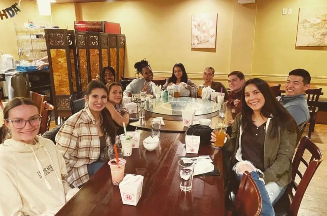 A group of casually dressed students sit at a restaurant with Chinese takeout boxes and boba tea in plastic cups with straws on the table in front of them.