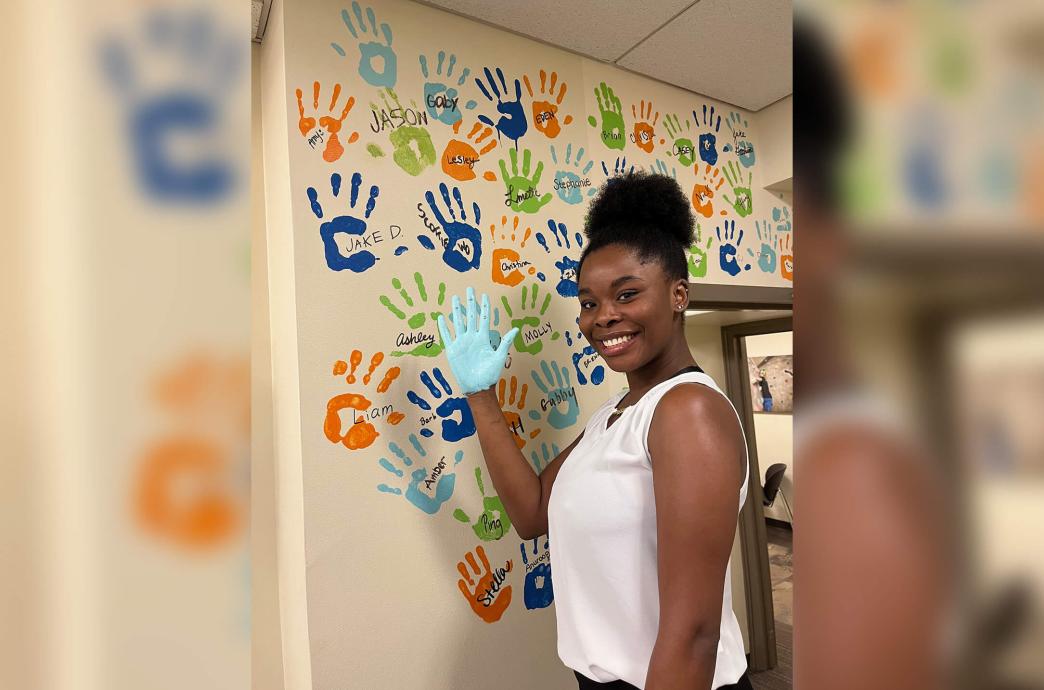 A female student holds up her hand covered in light blue paint in front of a wall of multicolored handprints with names.