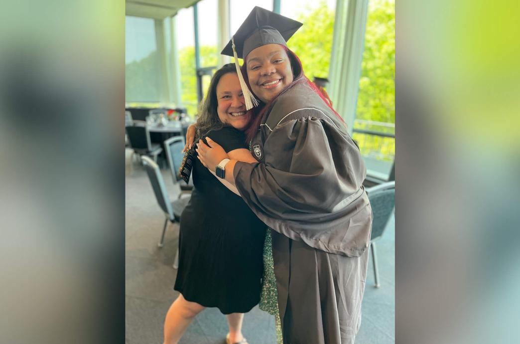 Carolina Hernandez side hugs a female student wearing a brown graduation cap and gown.