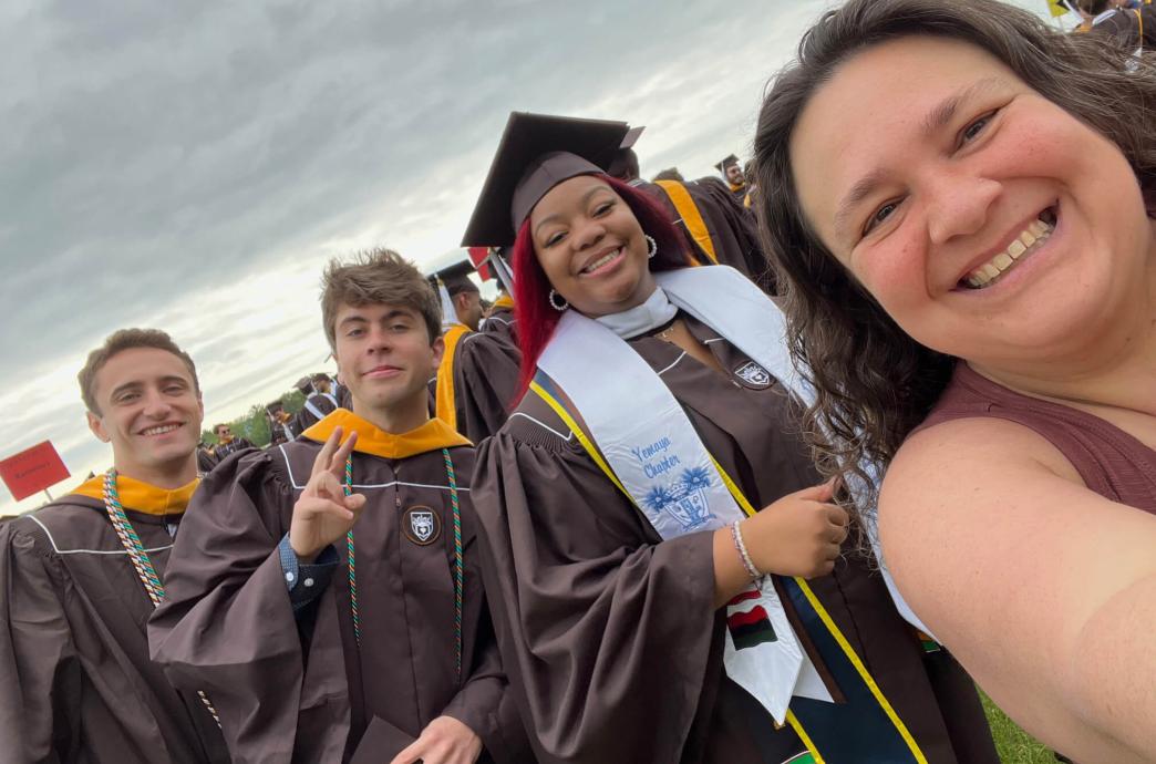 Carolina Hernandez takes a selfie of herself and 3 students wearing brown graduation gowns.