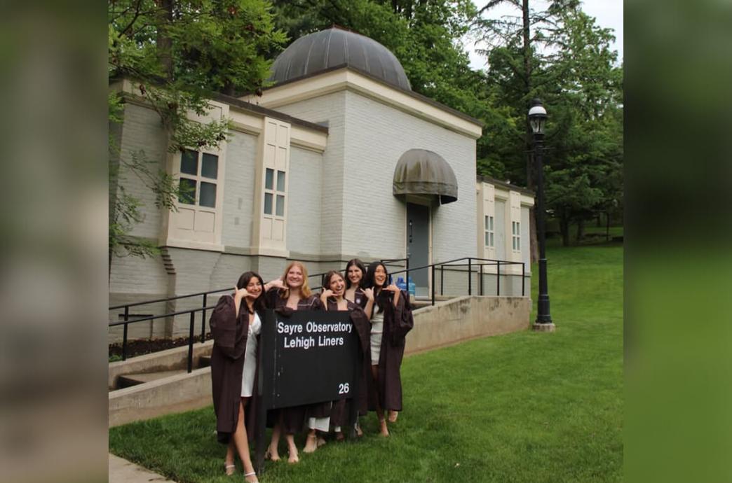 In brown graduation robes, 5 women stand with the “Sayre Observatory - Lehigh Liners” sign, holding their thumb and pinky fingers to their ear like a telephone.