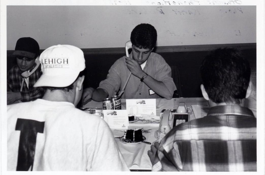 Students sit talking into phone receivers with plates of chocolate cake and cans of birch beer in front of them, with the focus on one student looking down.