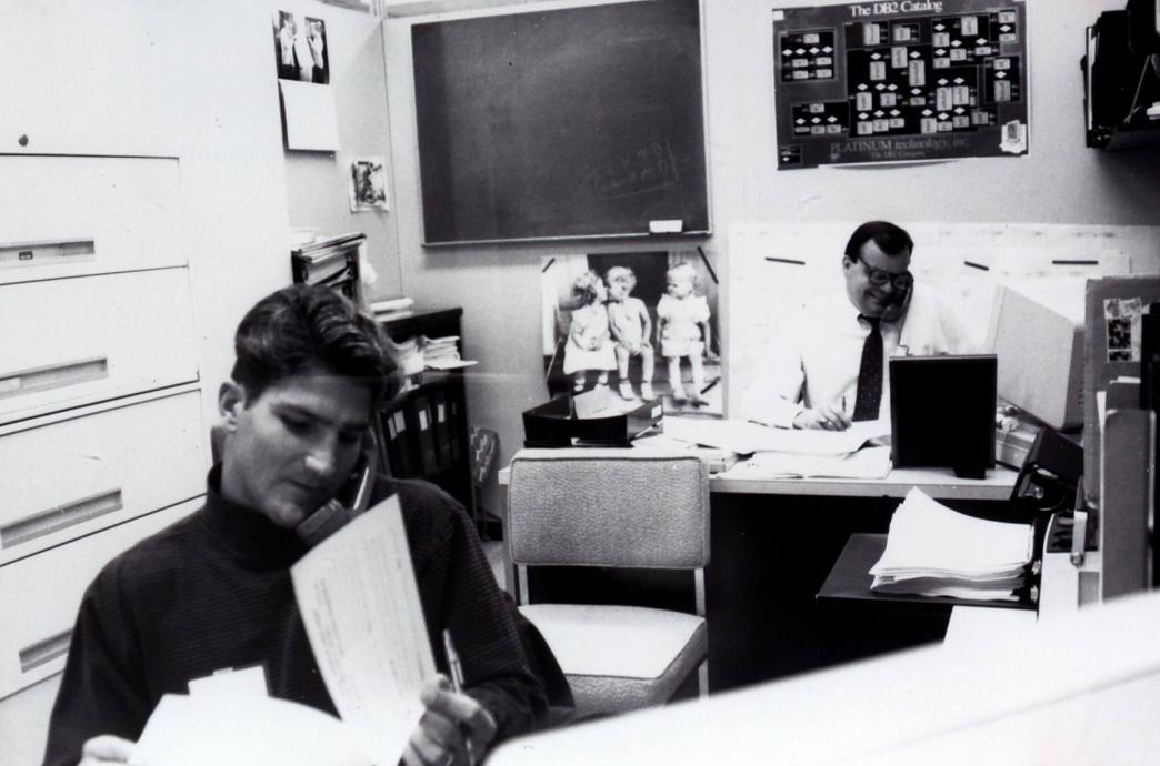 Two alumni sit in a small office while talking with phone receivers in the crooks of their necks and flipping through papers with pens in their hands.
