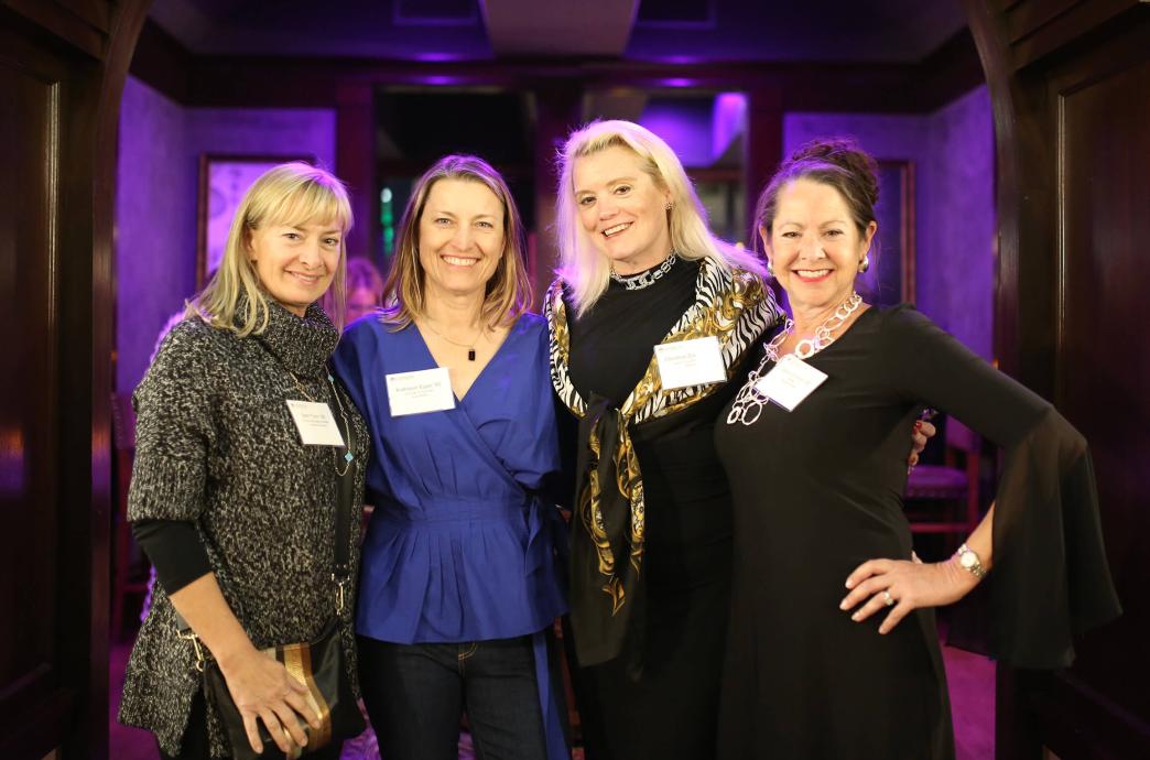 Four alumnae stand shoulder-to-shoulder for a photo, smiling and wearing professional attire and accessories.