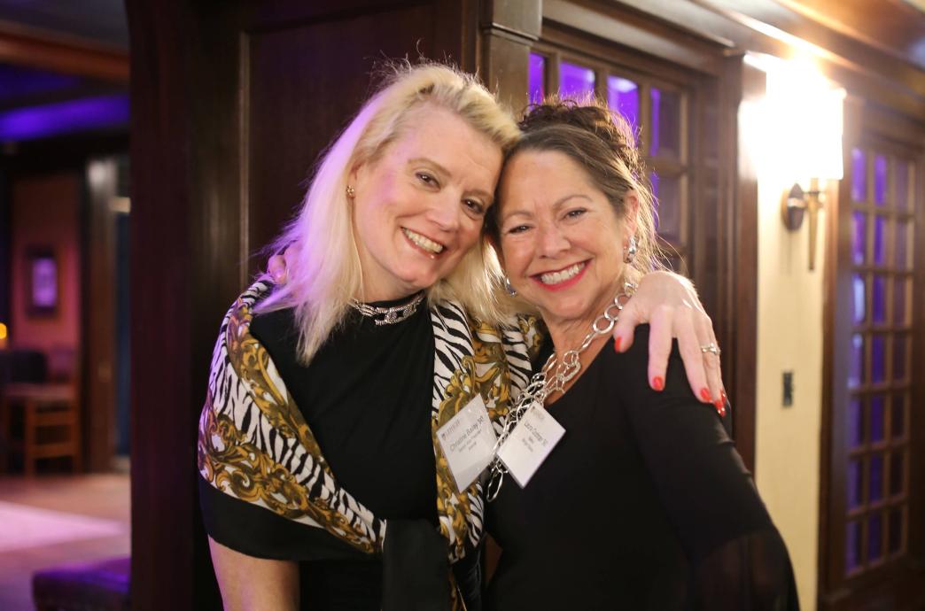  A blonde woman puts her arm around another and they lean theri heads together, smiling for a photo.