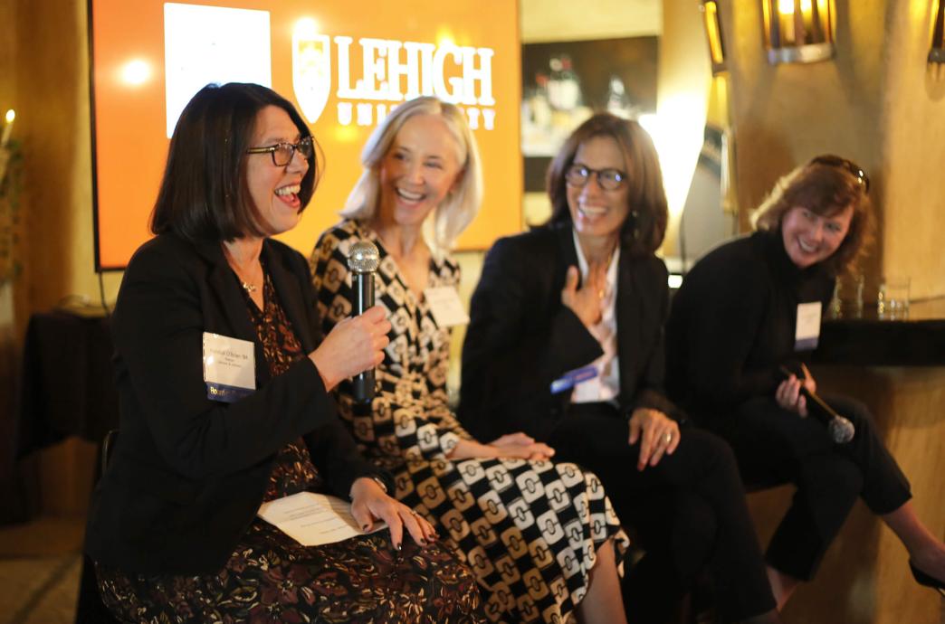 Four female panelists sit on bar height stools, the first speaking with a microphone while the others look on, smiling and laughing.