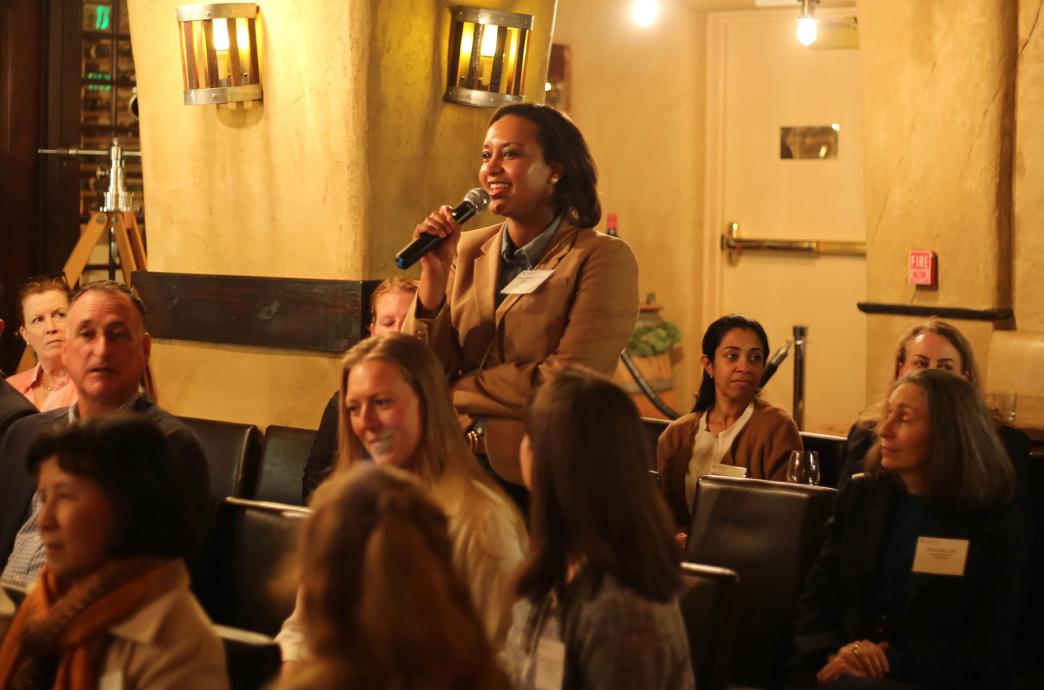 An alumna wearing a dark tan blazer stands at her audience seat holds a microphone and smiles while speaking in a warm lit room.