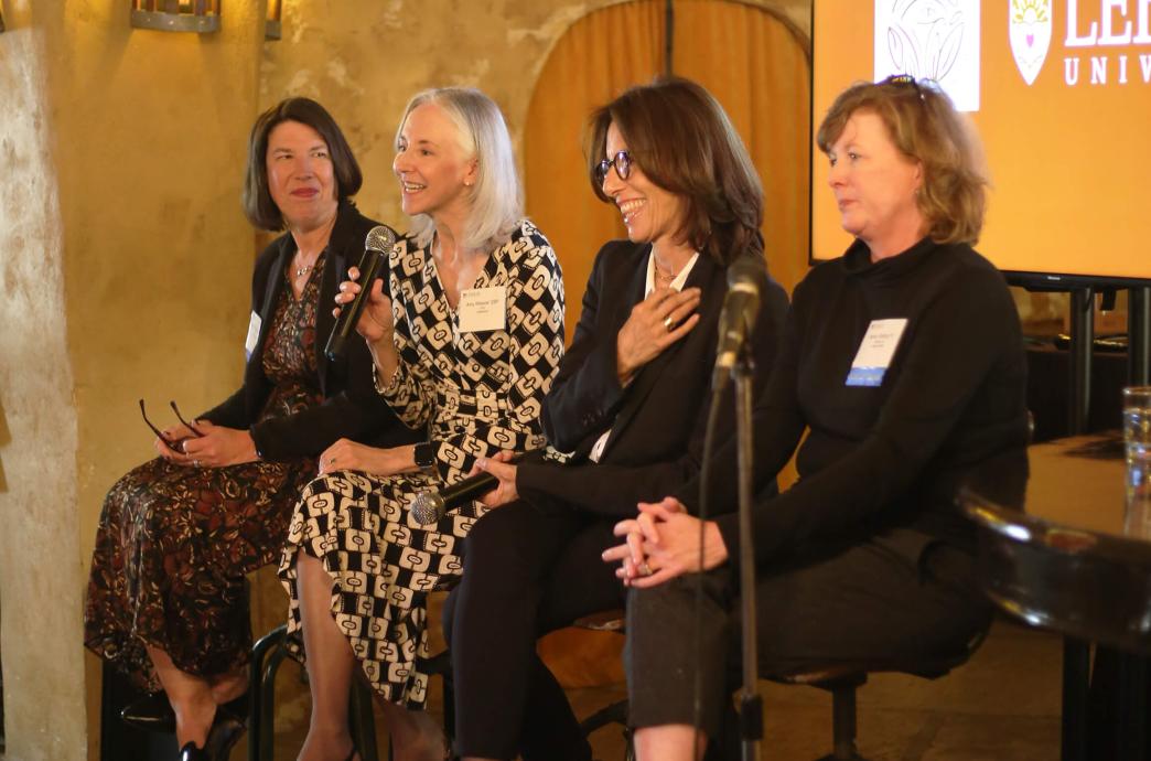 Four female panelists sit on bar height stools, the second speaking with a microphone, the one to her right smiling with her hand on her chest.