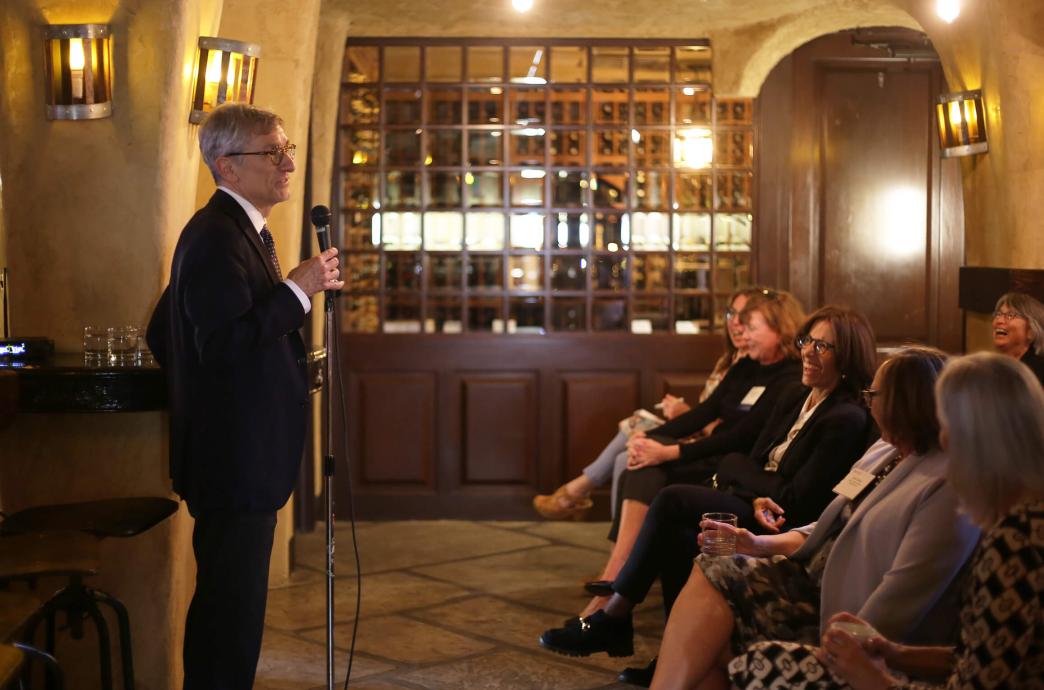 President Joe Helble stands with a microphone in front of an audience of women sitting with crossed legs in a warm lit room.