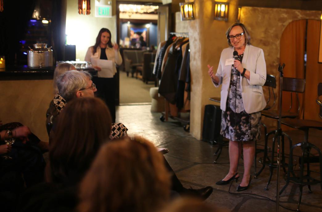 Beth Dolan stands in front of the attendees with a microphone in her right hand while gesturing with her left.