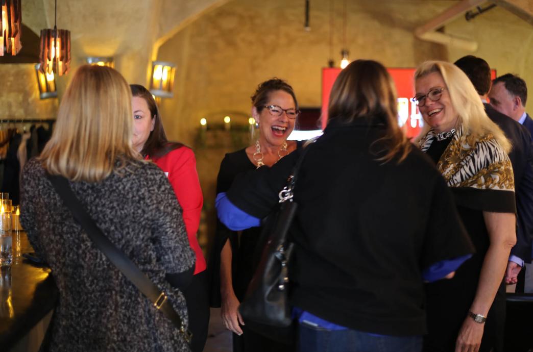Five alumnae stand socializing, smiling and laughing while wearing professional attire and statement accessories.