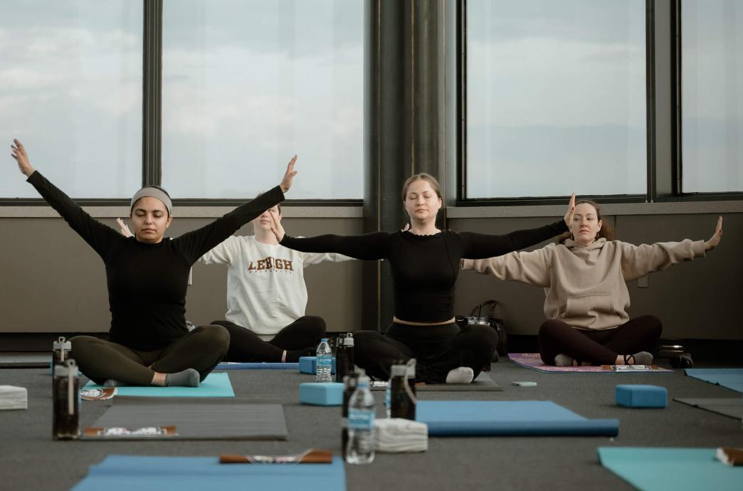 During a yoga session in the observation tower room of Iacocca Hall, 4 women sit on the floor with eyes closed, legs crossed, and arms outstretched.