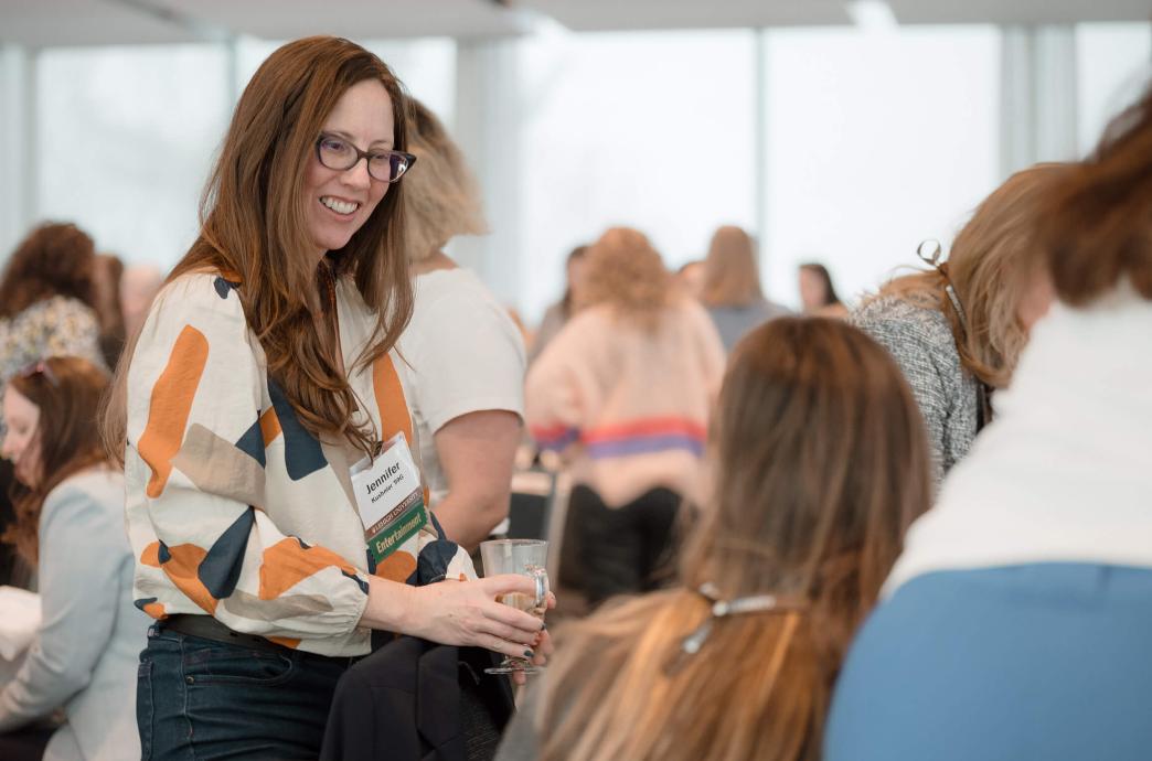 A woman wearing glasses and a blouse walks around a conference room, stopping at a table to chat with an attendee.