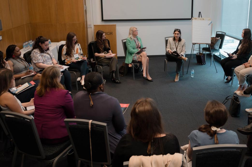 In a medium sized room, a large group of women sit in a circle of chairs with one woman leading from her chair next to an easel.