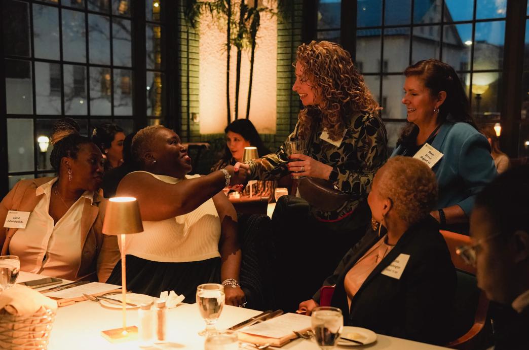 A table of women sitting with drinks in front of them turn to greet two other women approaching them and the center seat shakes hands with one.
