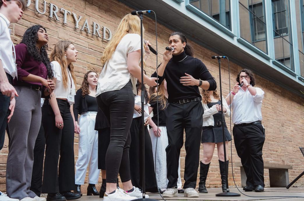 In an outdoor patio at Iacocca Hall, an a capella group sings into microphones, with a man and woman taking the leads facing each other.