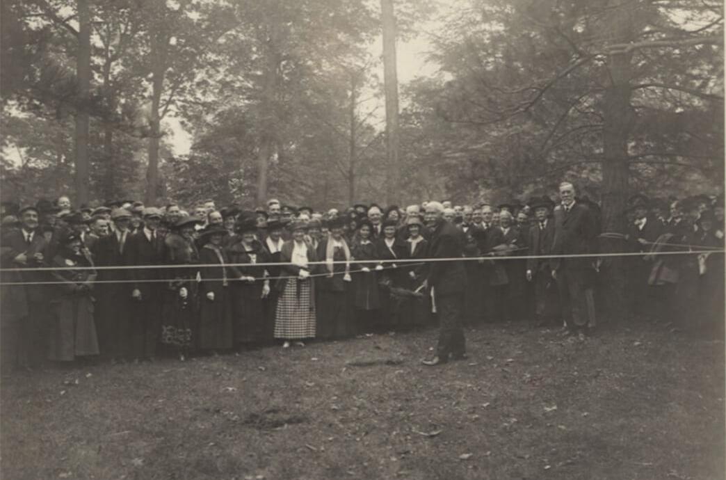 A black and white photo of a crowd of people gathering around a man holding a shovel.